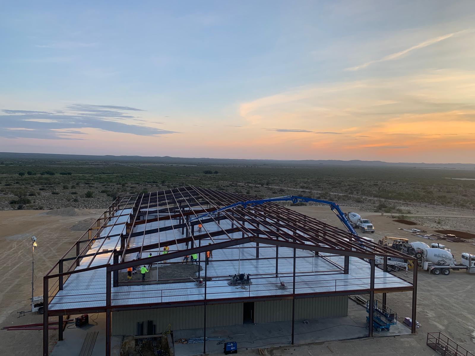 Metal building frame under construction at sunset with workers and concrete trucks